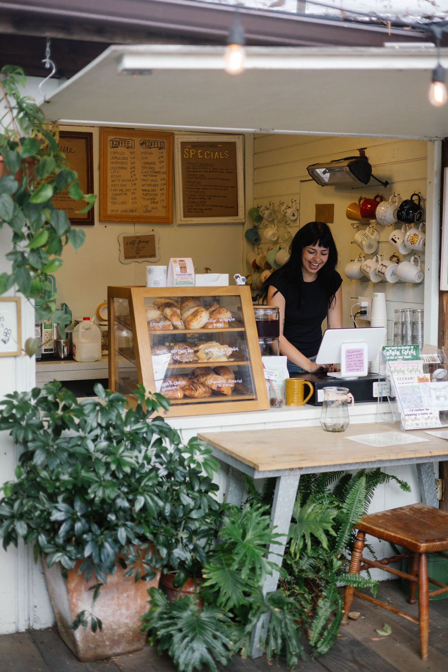 Smiling woman working at a charming outdoor cafe counter with pastry display, plants, and hanging mugs.