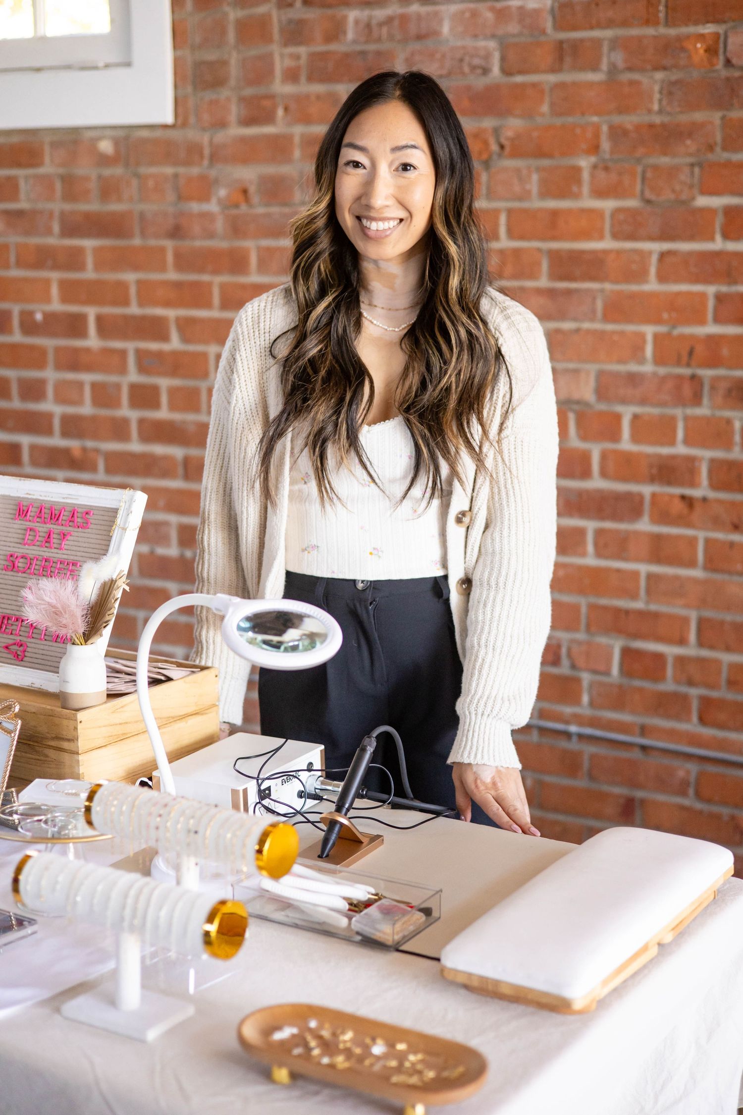 Smiling woman with long dark hair stands at a craft jewelry table with tools in front of a brick wall.