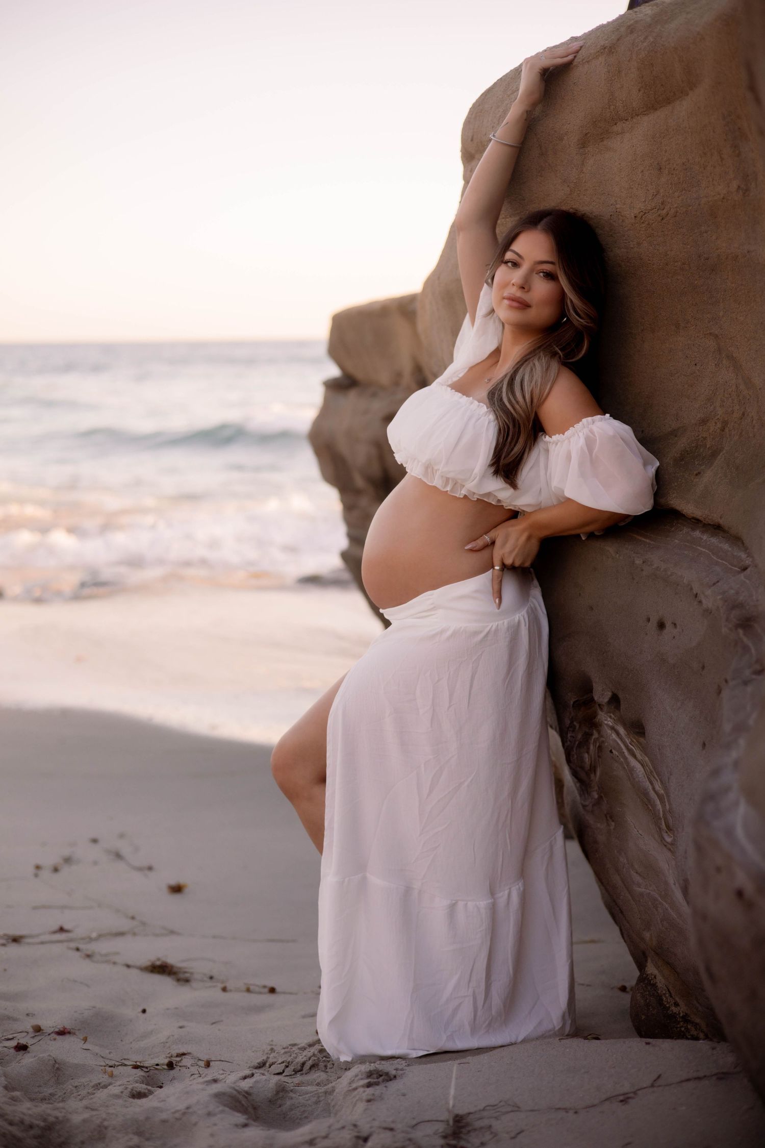 Pregnant woman in white off-shoulder dress leaning against beach rock at golden sunset.