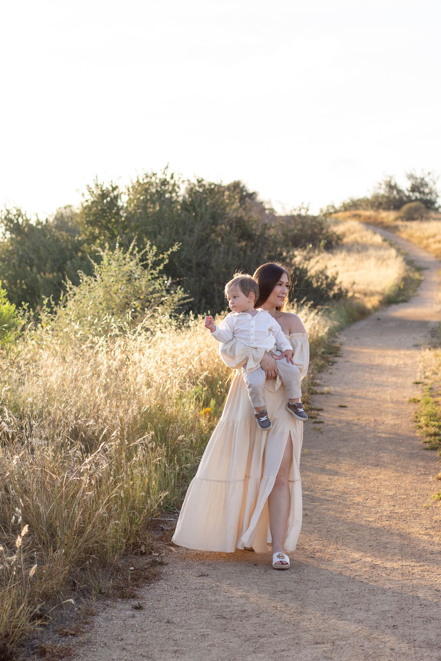 Mother in elegant cream gown carries toddler along sunny dirt path surrounded by golden grass.