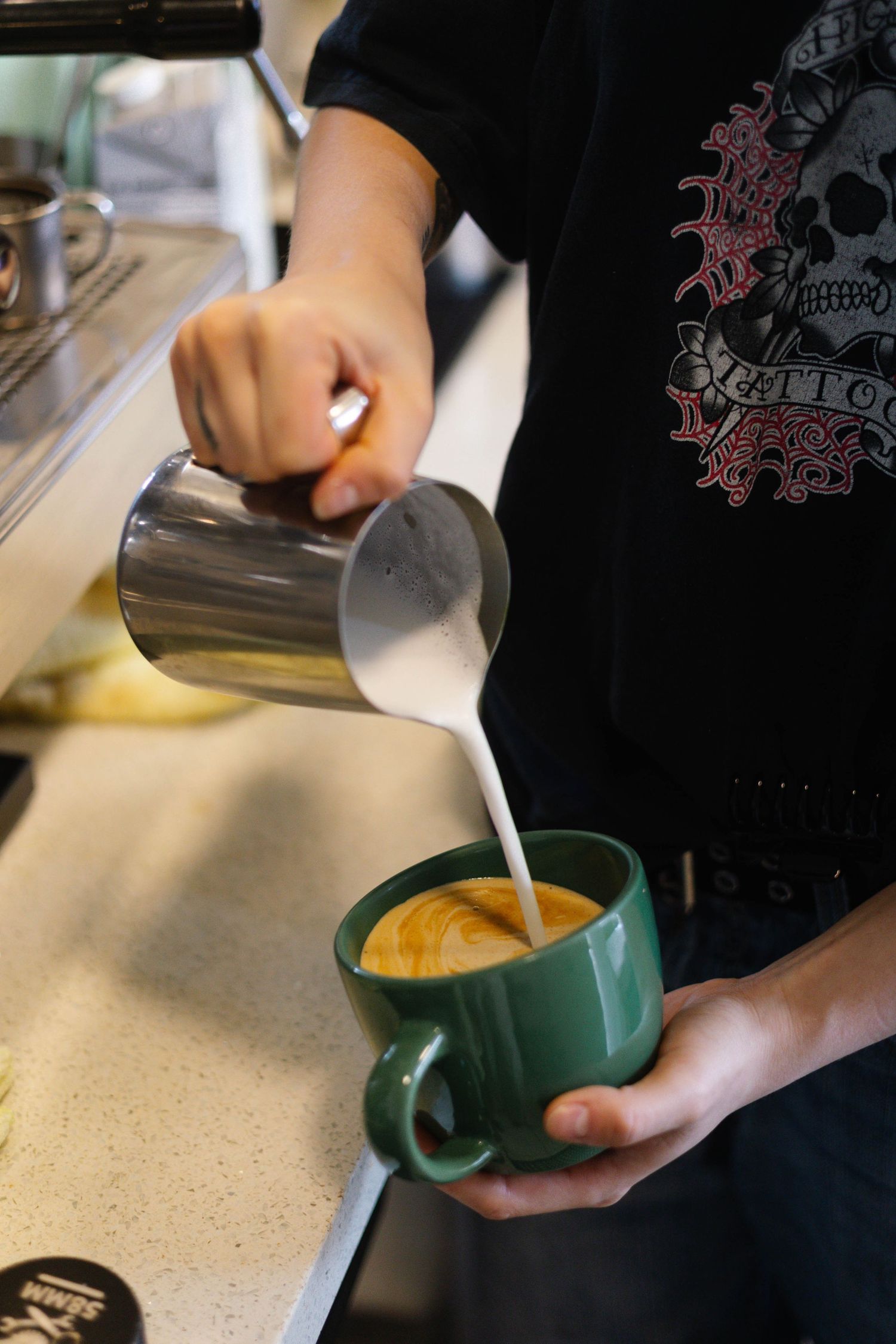 Barista pouring steamed milk into a green ceramic mug to create a latte in a coffee shop.