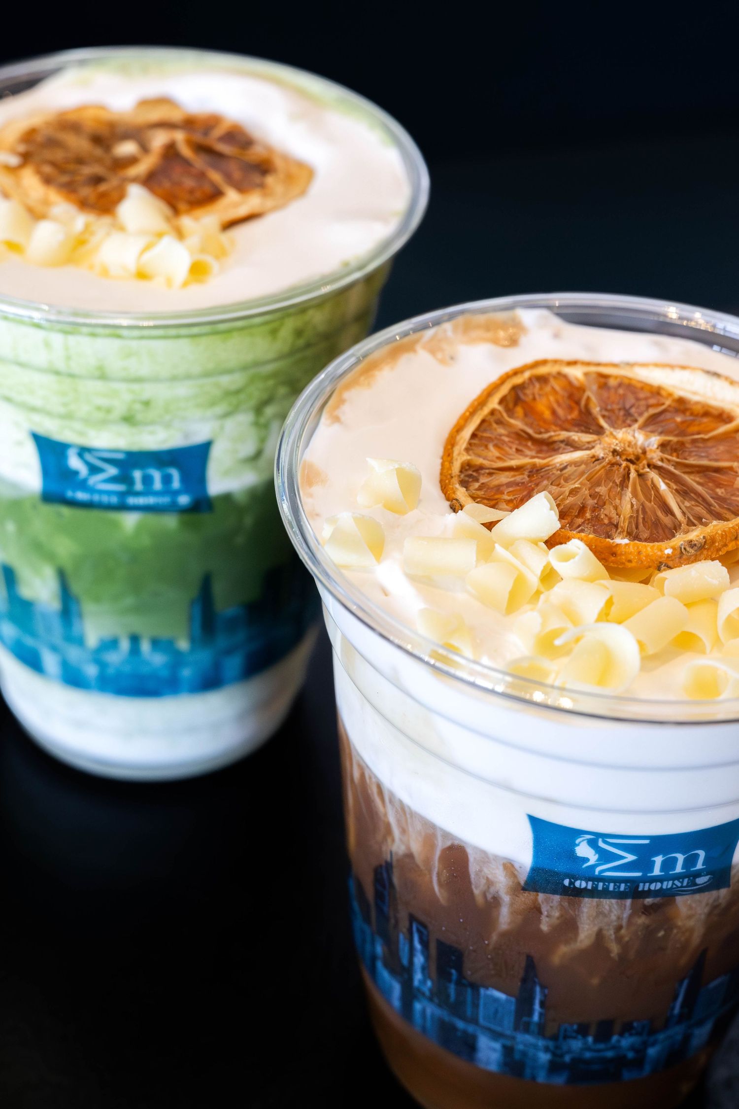 Two specialty coffee drinks in branded cups topped with dried orange slices and white chocolate shavings on a dark background.