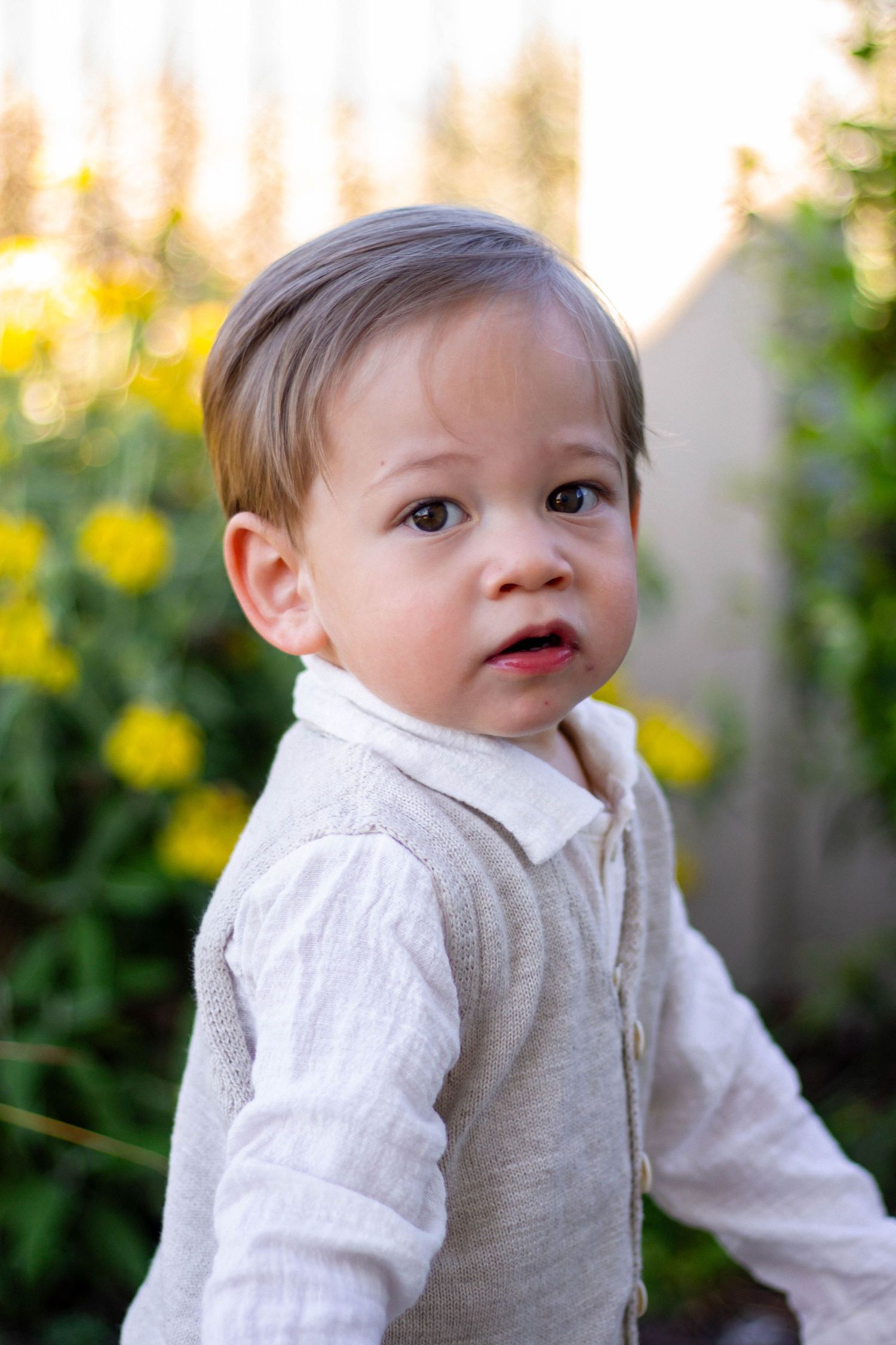 A cute toddler in a beige outfit stands outdoors with yellow flowers in the background.