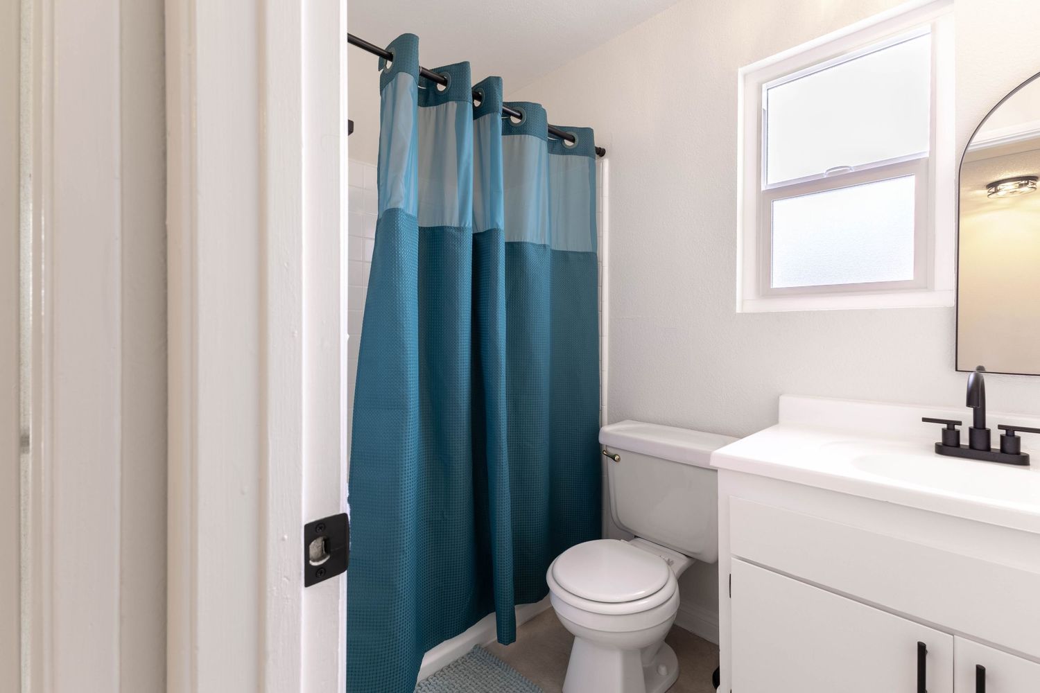 Clean bathroom with teal shower curtain, white vanity, toilet, and natural light from a small window.