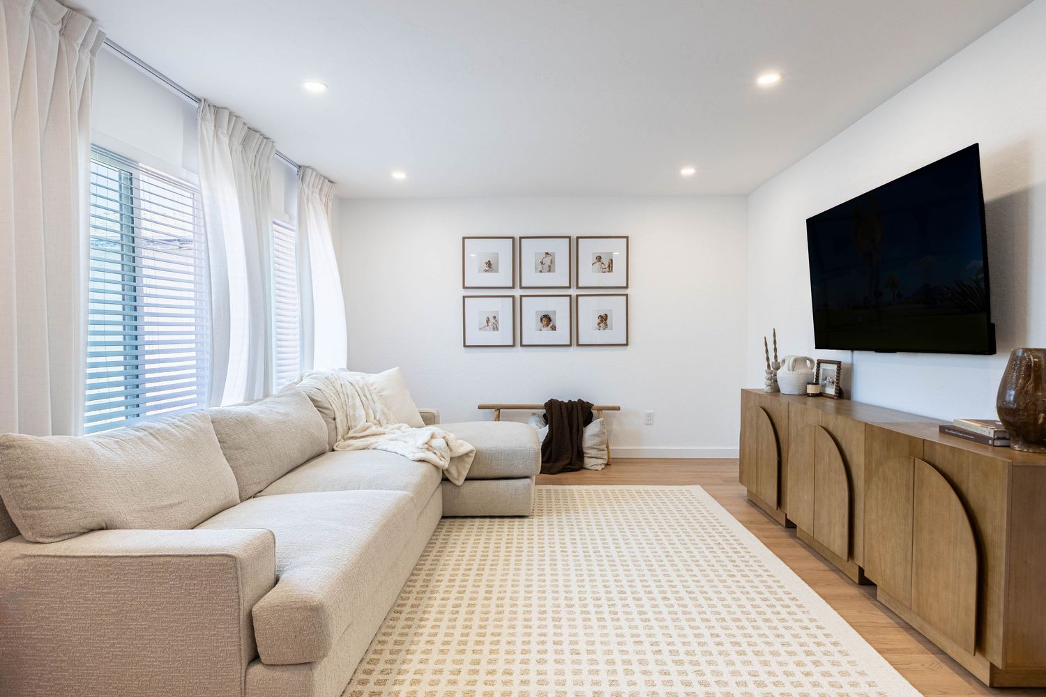 Modern living room with beige sectional sofa, wall-mounted TV, wooden sideboard, and neutral area rug on light hardwood floors.