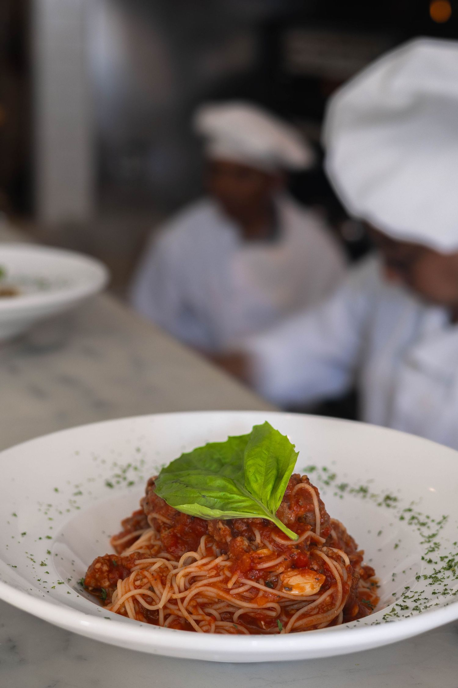 Spaghetti bolognese with fresh basil garnish on white plate, chefs in white uniforms working in background.