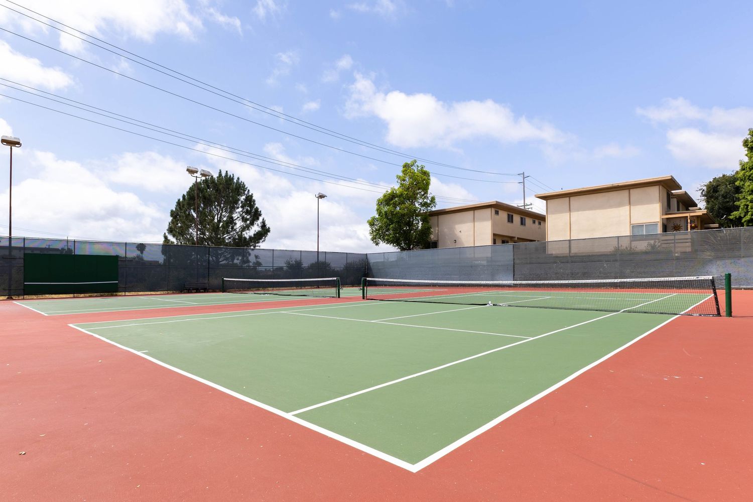 Outdoor tennis court with green and red surfaces, net, light poles, trees, and a building under a partly cloudy sky.