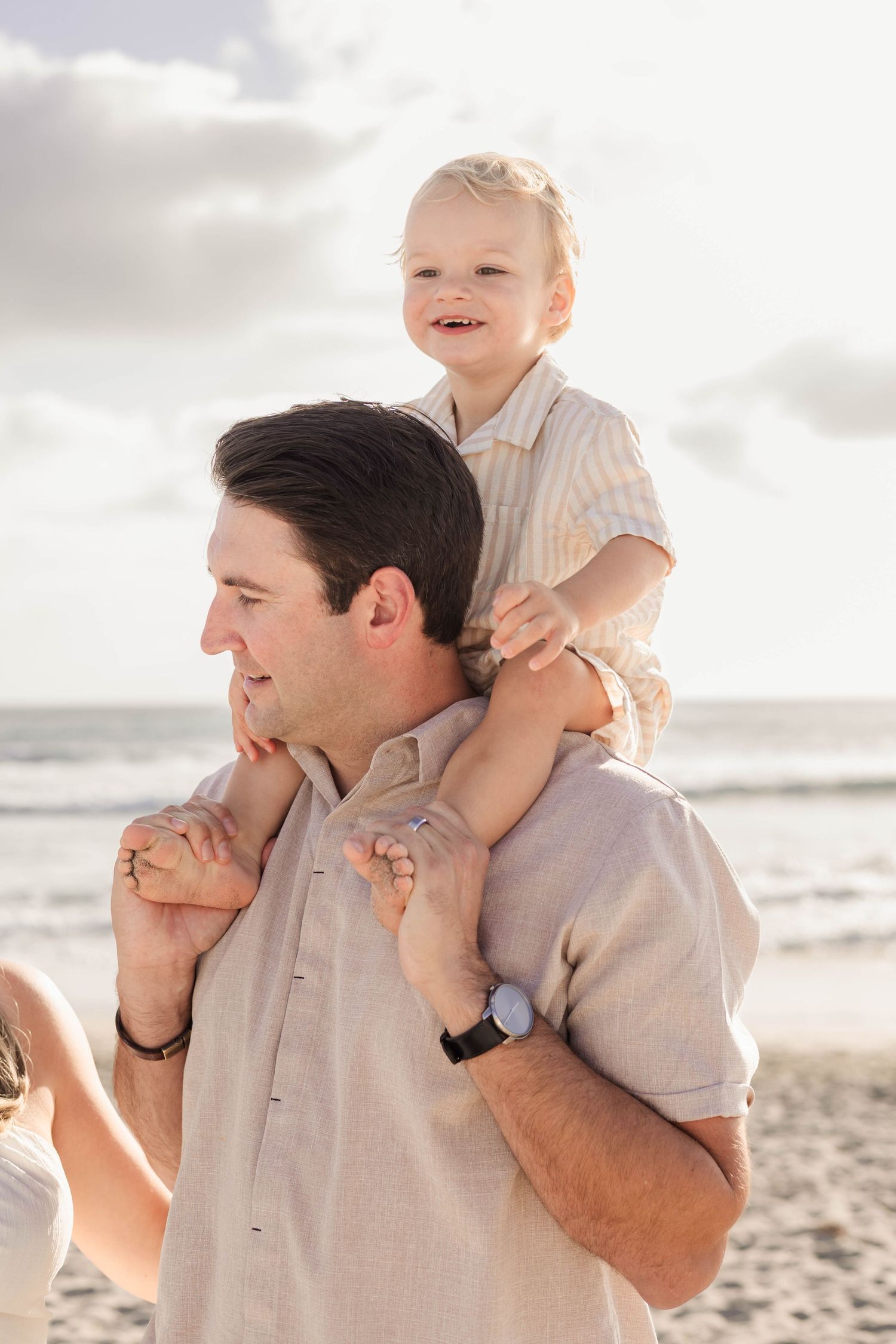 A smiling toddler sits on a man's shoulders at the beach, both wearing neutral linen outfits on a sunny day.