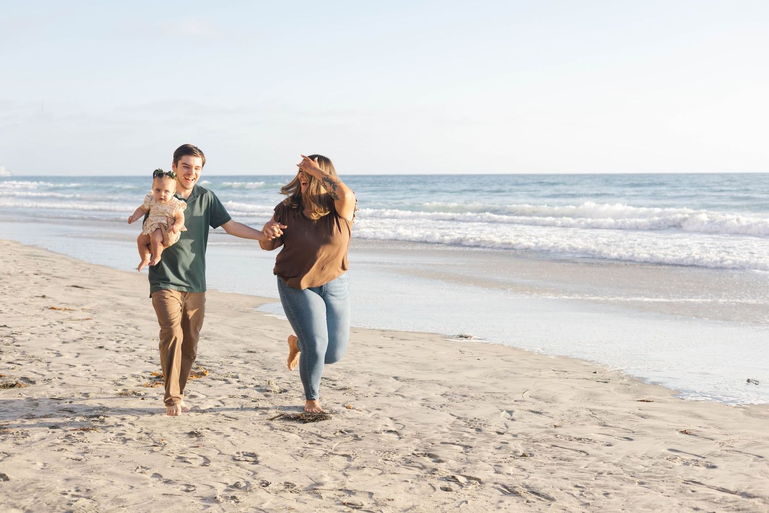 Happy young family with baby running on sandy beach near ocean waves on a sunny day.