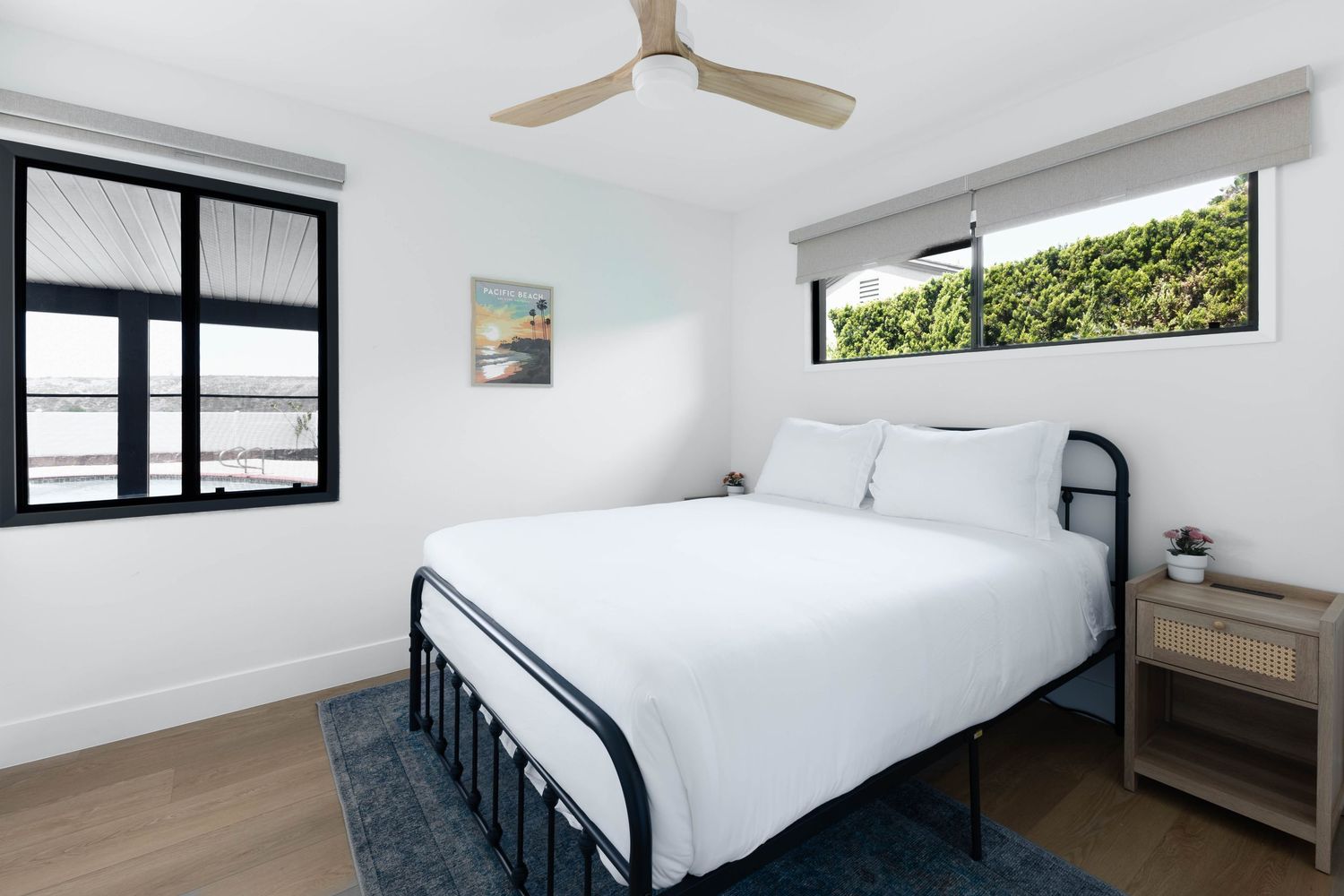 Modern bedroom with black metal bed frame, white bedding, hardwood floors, ceiling fan, and bright windows with garden views.