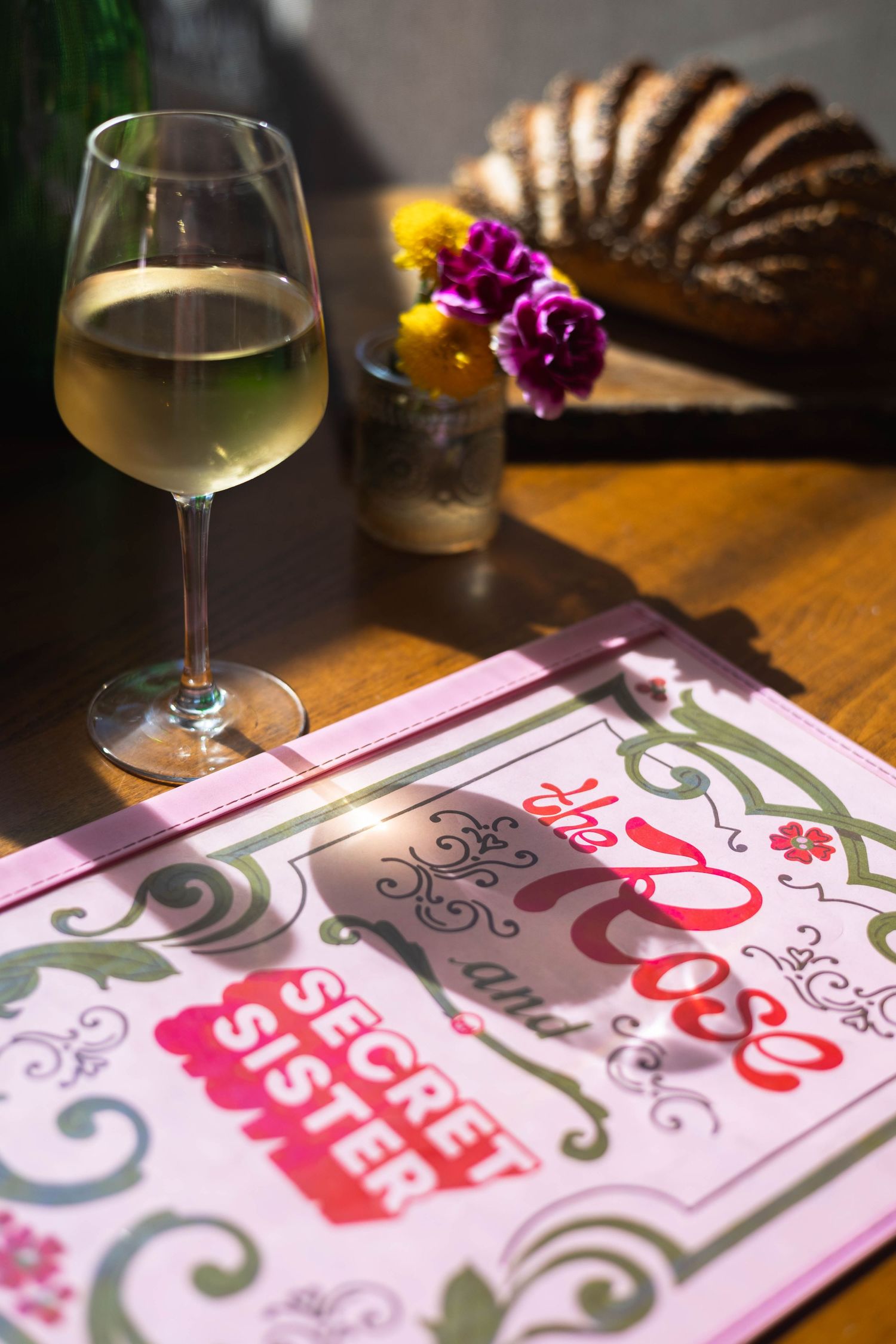 White wine glass beside The Rose and Secret Sister menu on wooden table with bread and purple flowers.