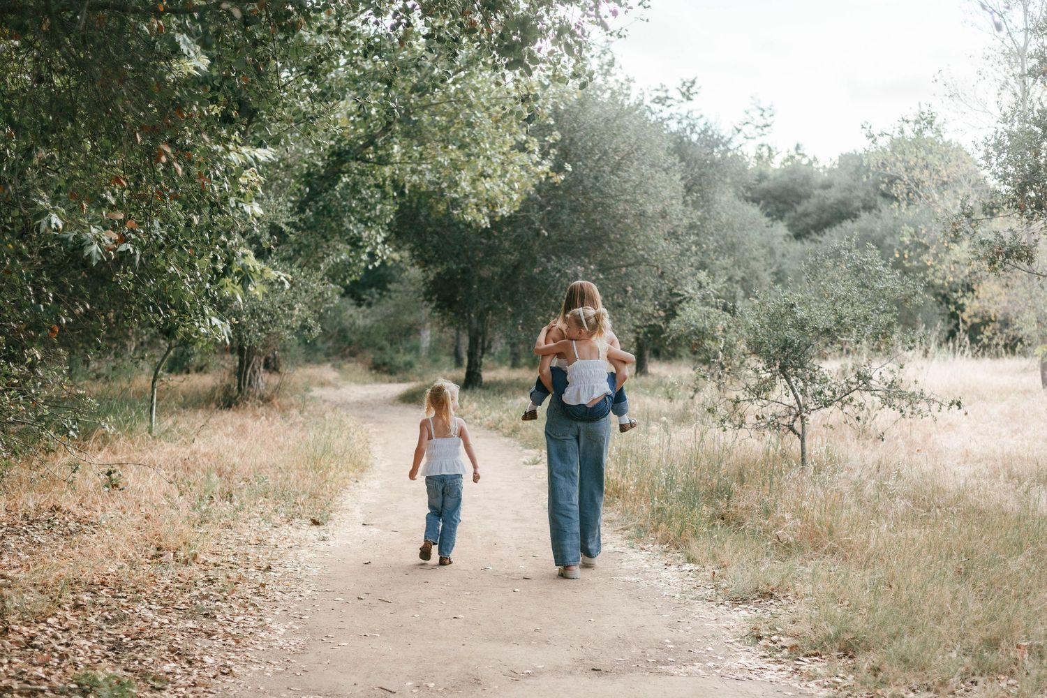 A mother walks with two young children along a serene dirt path through a lush green forest.