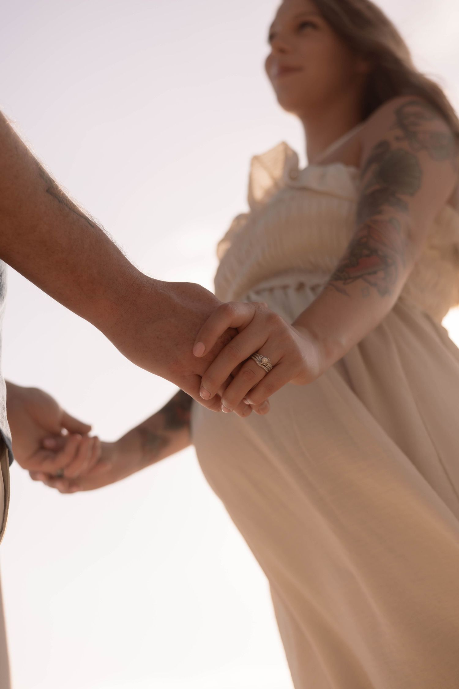 Pregnant woman in white dress with tattoos holds hands with tattooed man in soft backlit romantic photo.