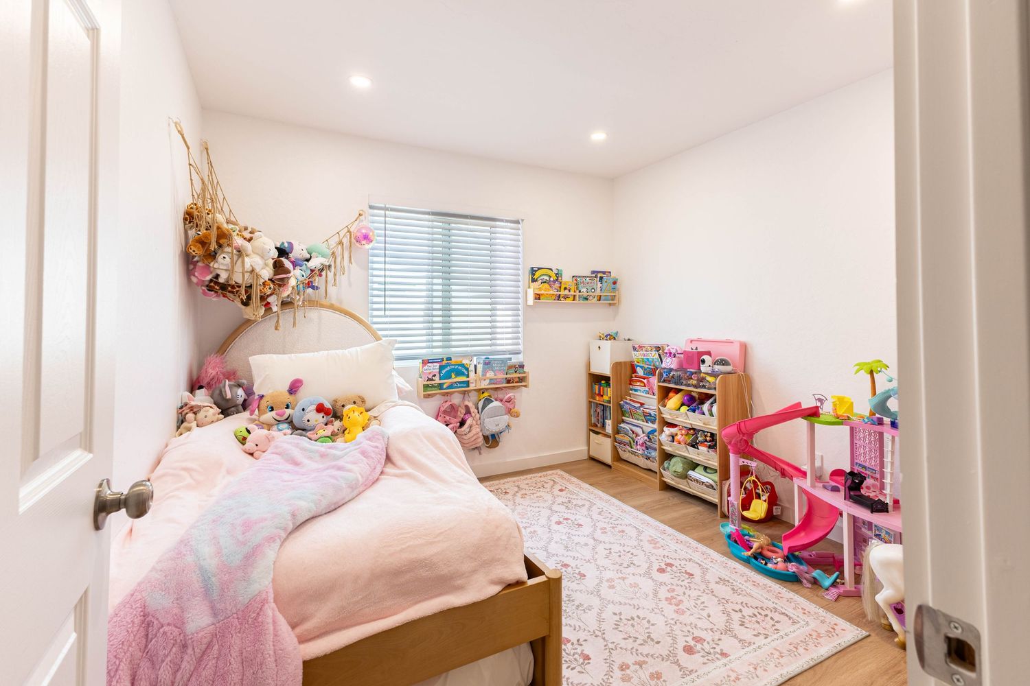 Bright girl's bedroom with pink bedding, toy storage shelves, and a dollhouse on hardwood floors.