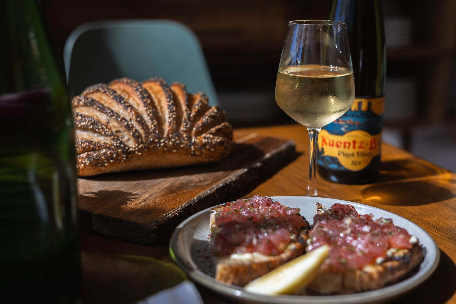 Rustic bread loaf on cutting board with charcuterie plate and white wine glass beside Riesling bottle on wooden table.