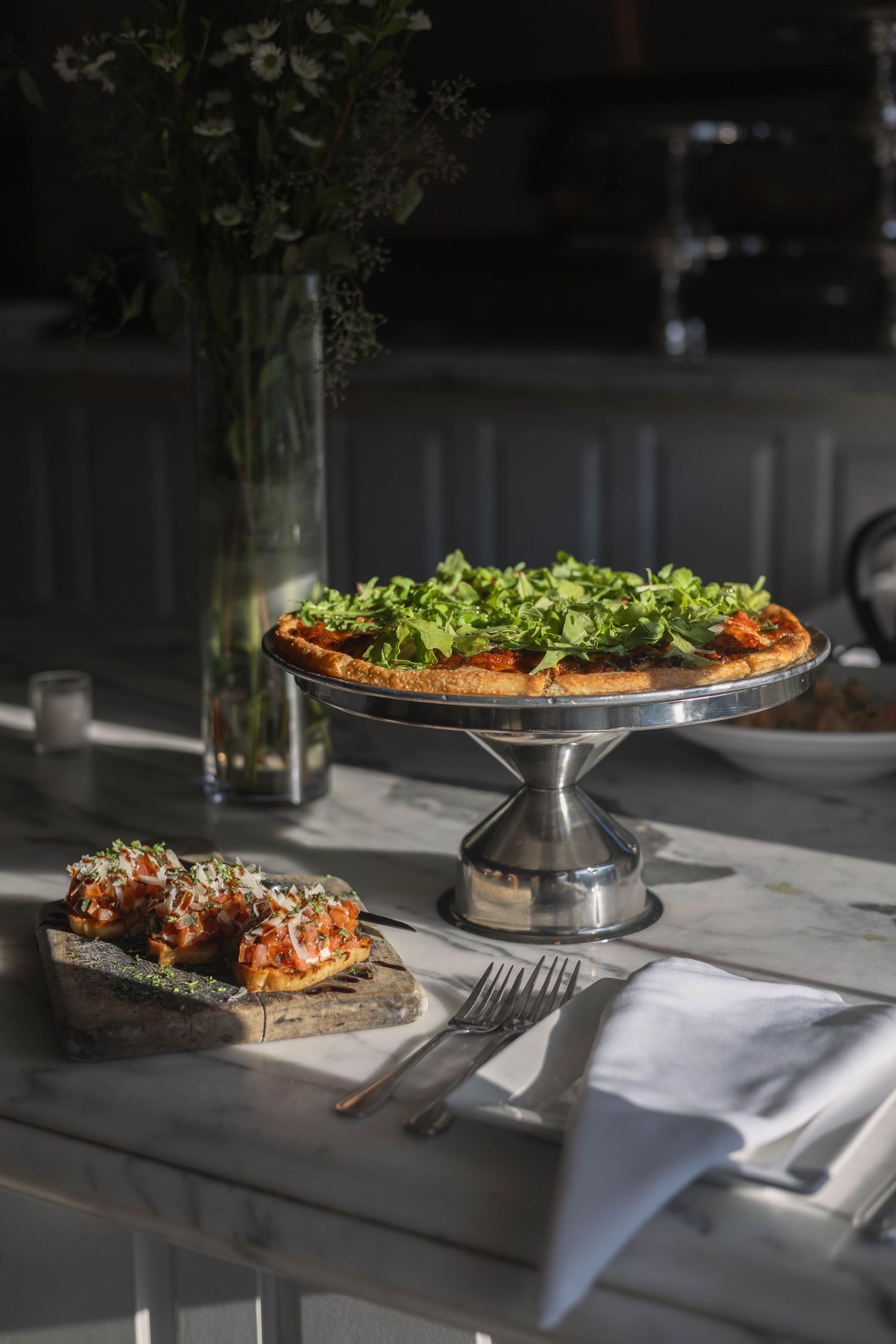 A gourmet pizza topped with fresh arugula on a silver cake stand, served on a marble table with bruschetta and cutlery.