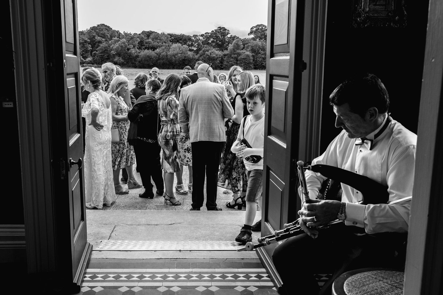 Black and white photo of wedding guests mingling while musician performs in doorway.
