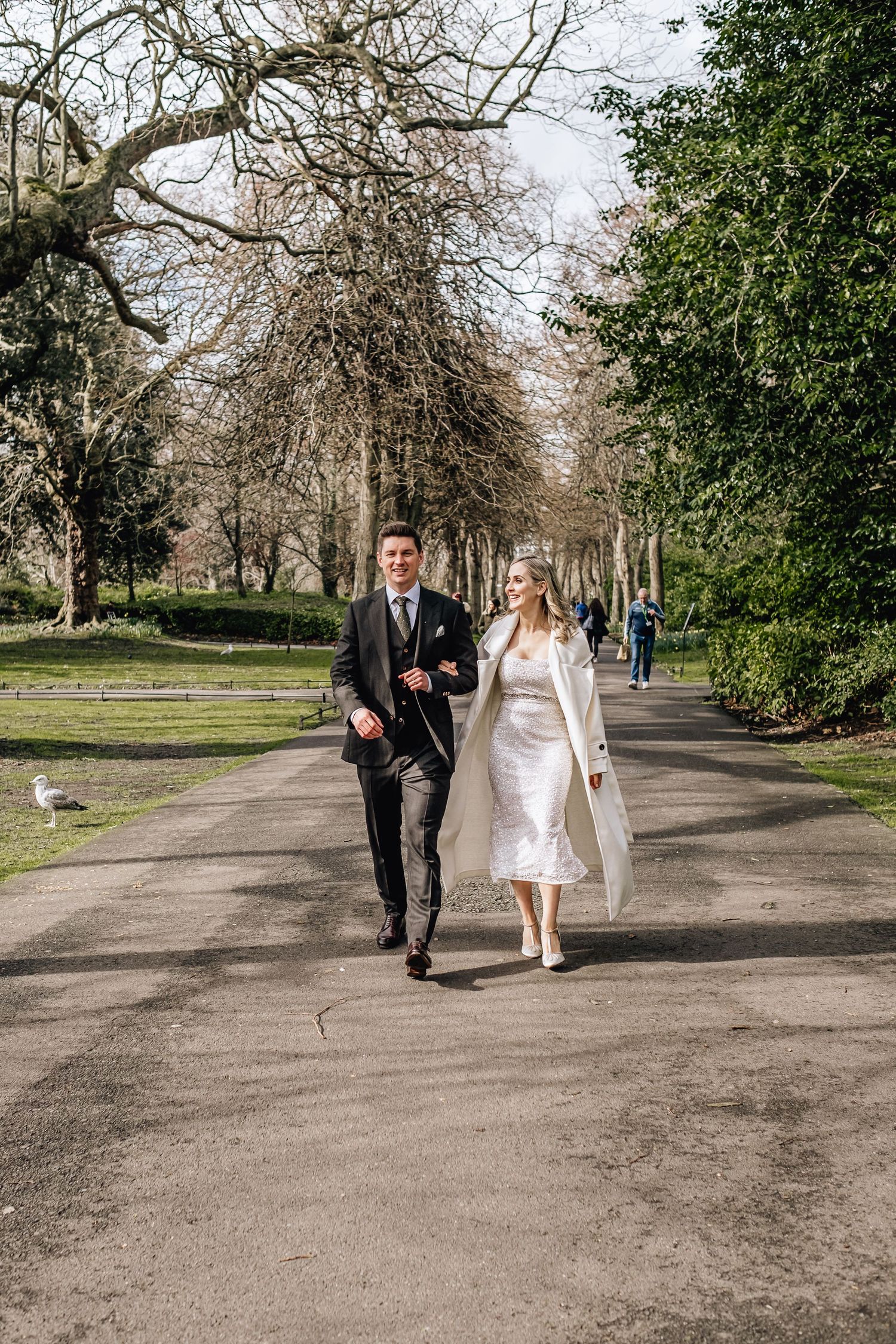 Couple walking along tree-lined park path in formal wedding attire.