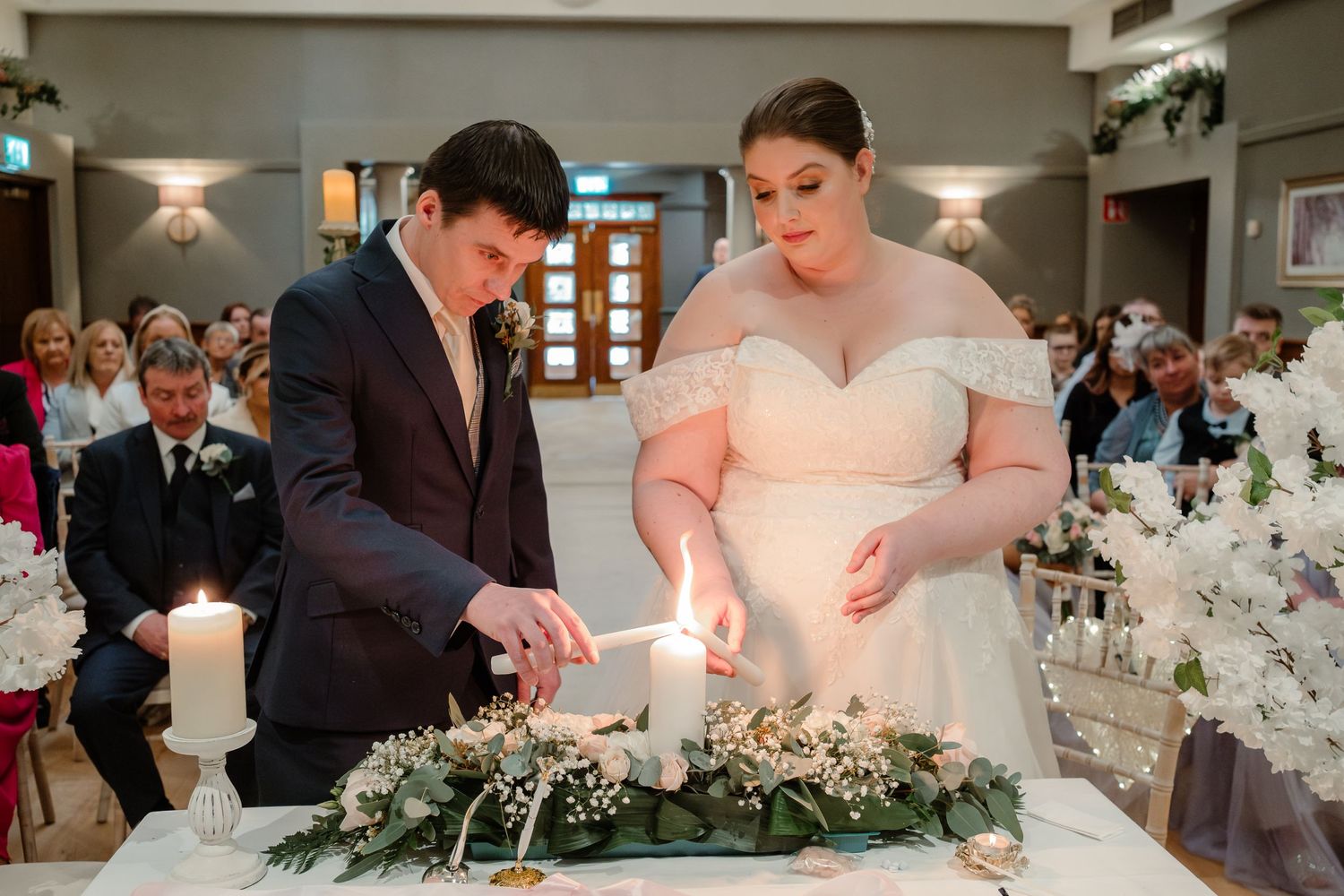 A couple performs a unity candle lighting ceremony at their wedding reception surrounded by guests.