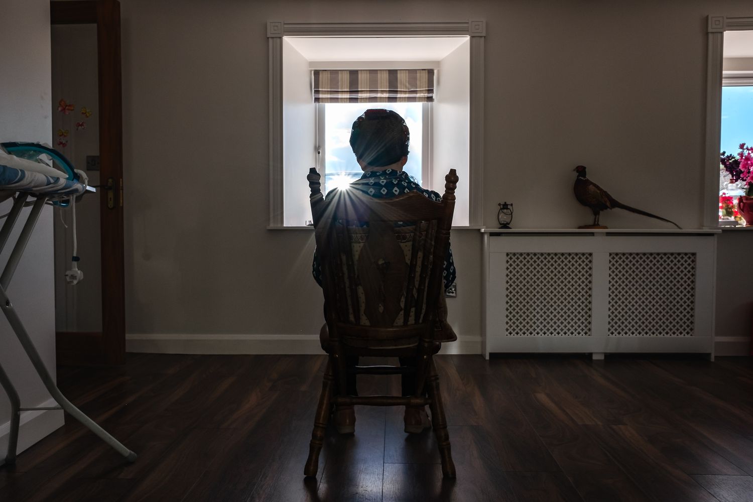 A silhouette sits in a chair facing a bright window in a minimalist room with hardwood floors.