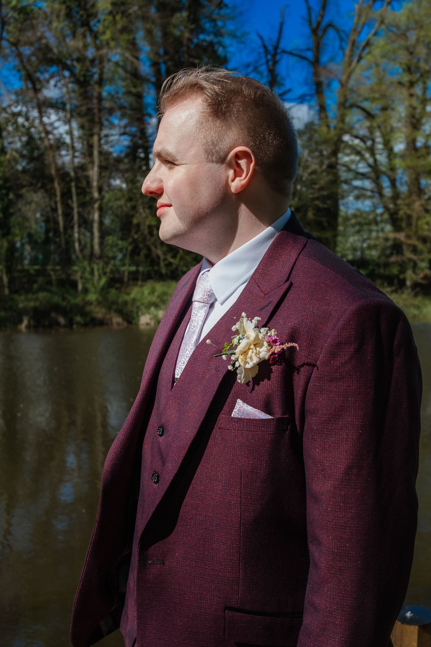 Men in burgundy suits stand beside a lake on a sunny day with trees in background.