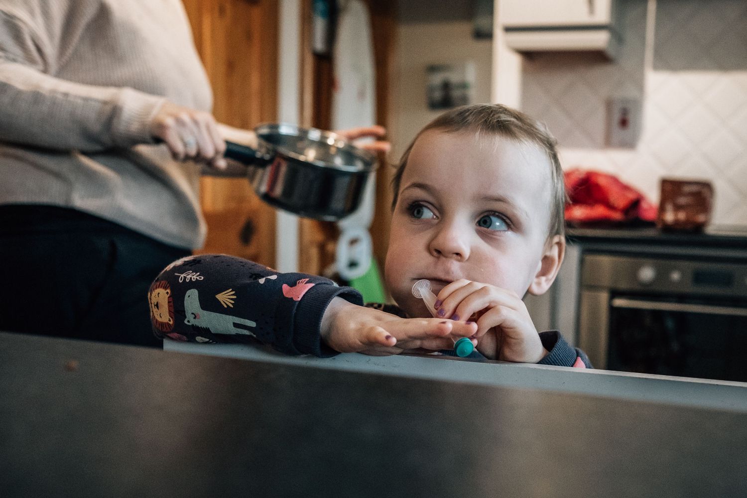 Child resting chin on dark counter while someone pours from a bottle in kitchen.