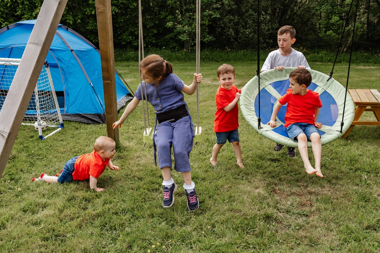 Kids playing in backyard with swings and blue tent on green grass.