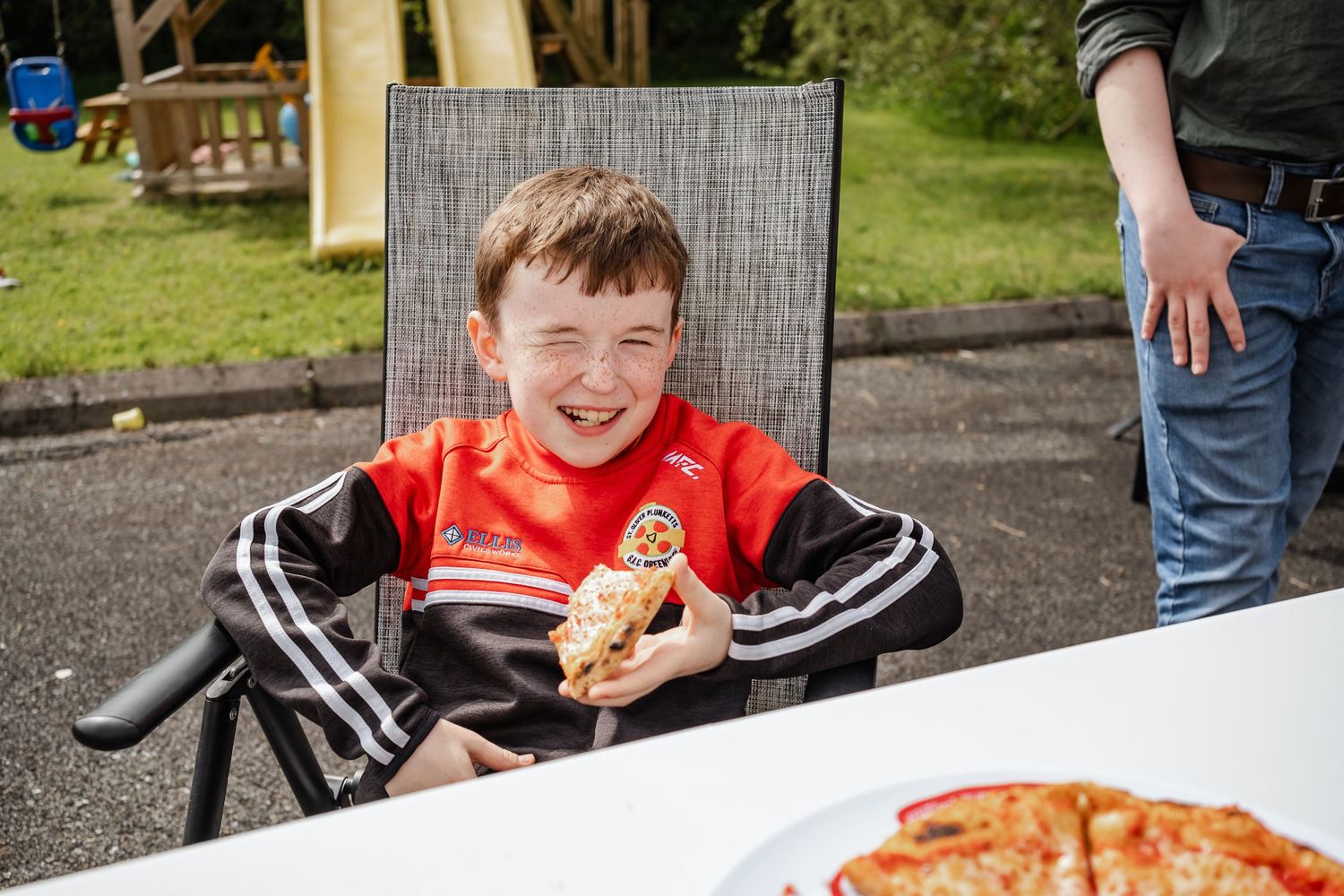 Happy child in red shirt sitting outdoors with pizza on the table.