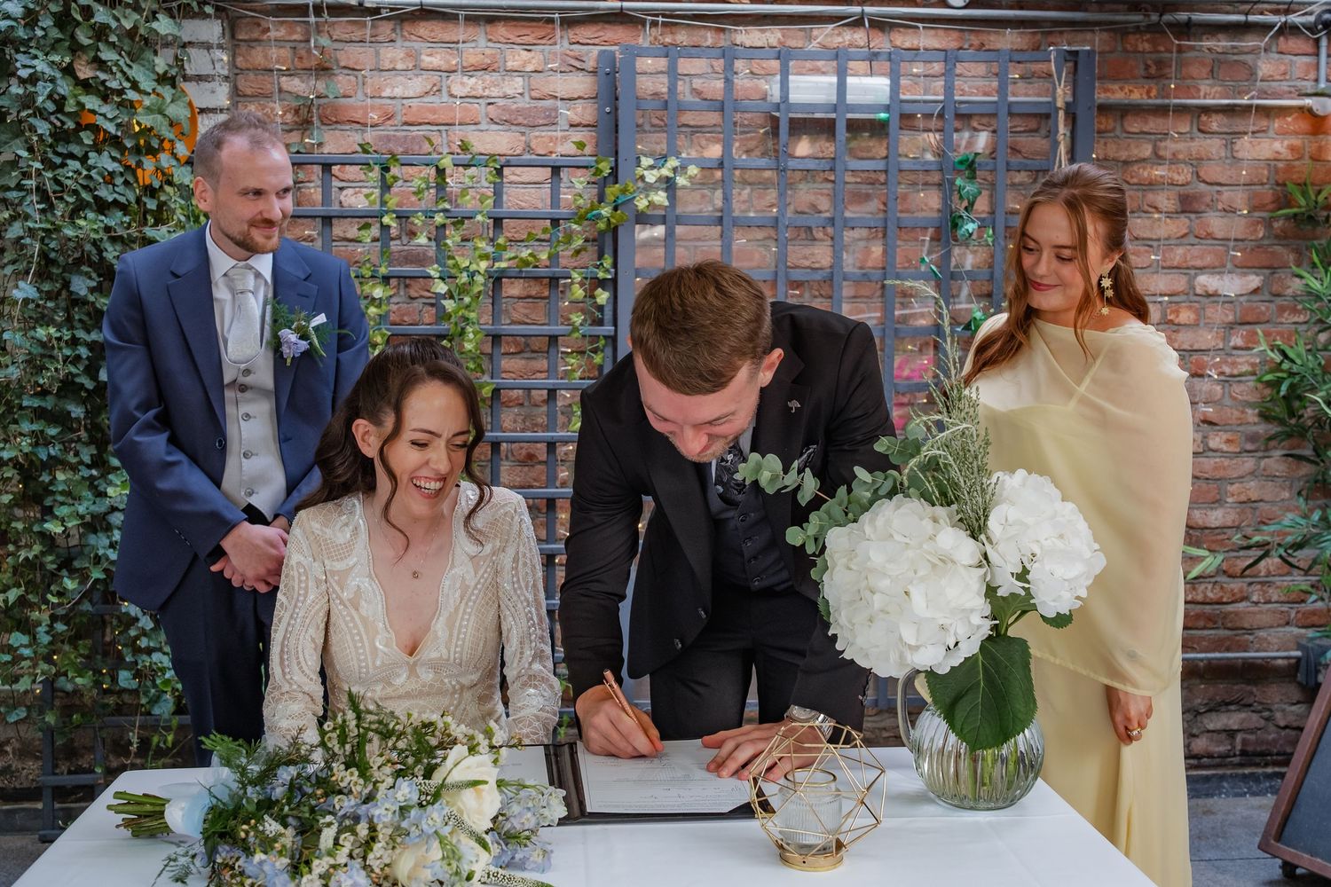 Wedding couple and guests gathered around a table signing documents with white floral arrangements at an outdoor venue.
