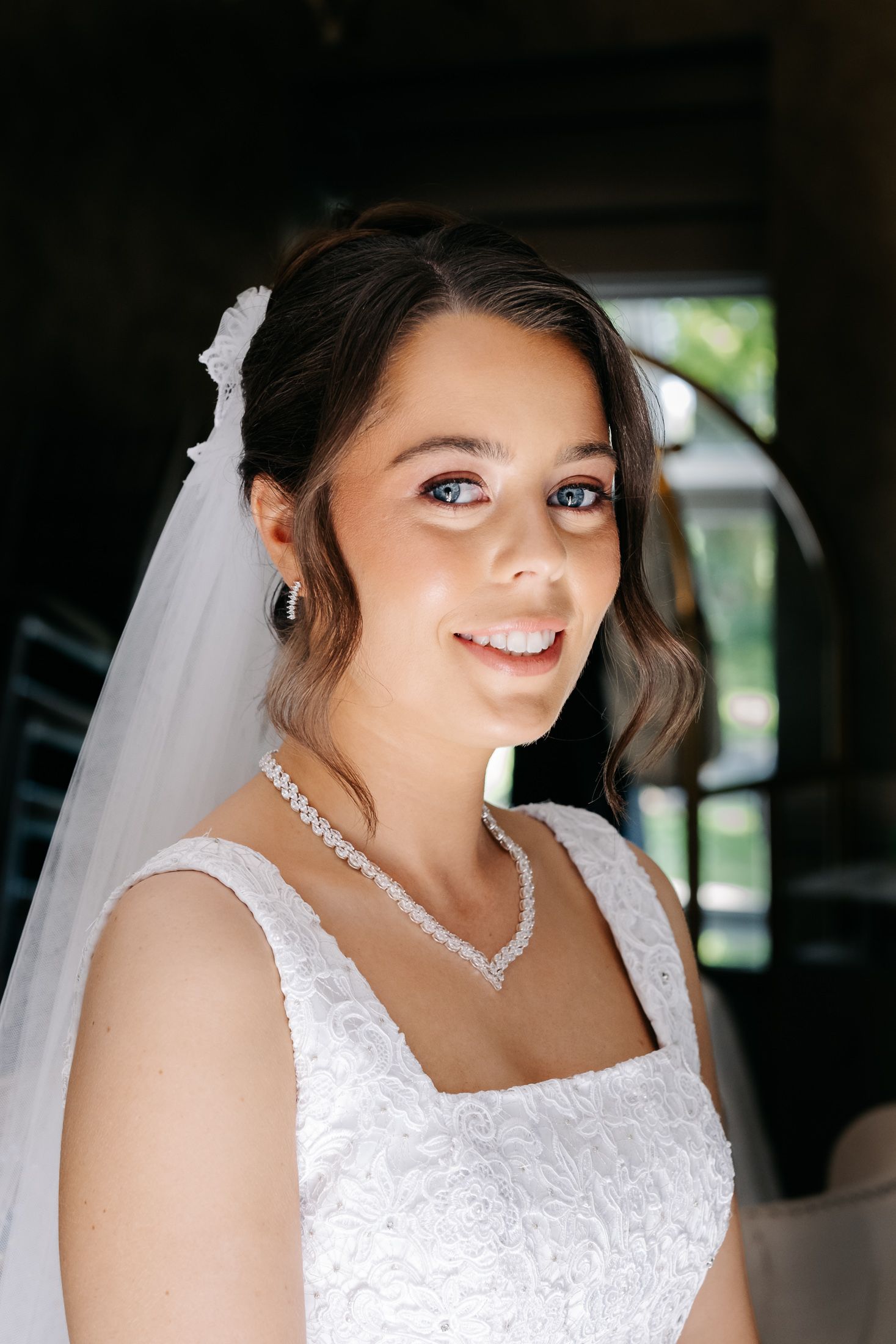 Smiling bride in white lace gown and veil with pearl necklace poses indoors on her wedding day.