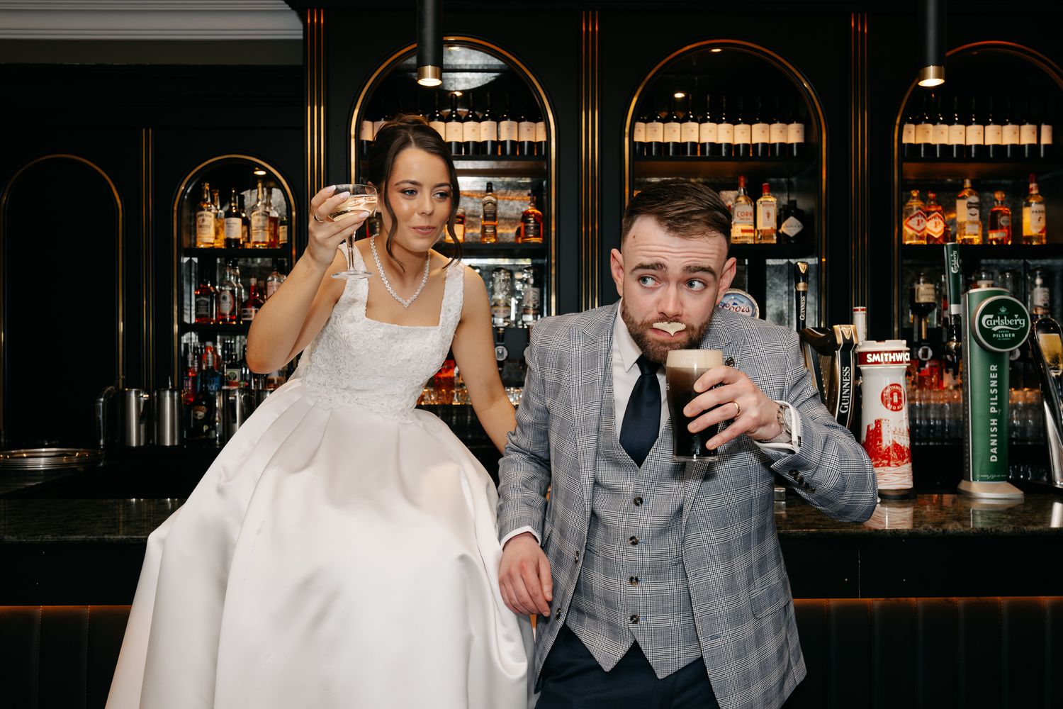 Bride and groom sitting at a bar, laughing and enjoying drinks, with shelves of bottles in the background.