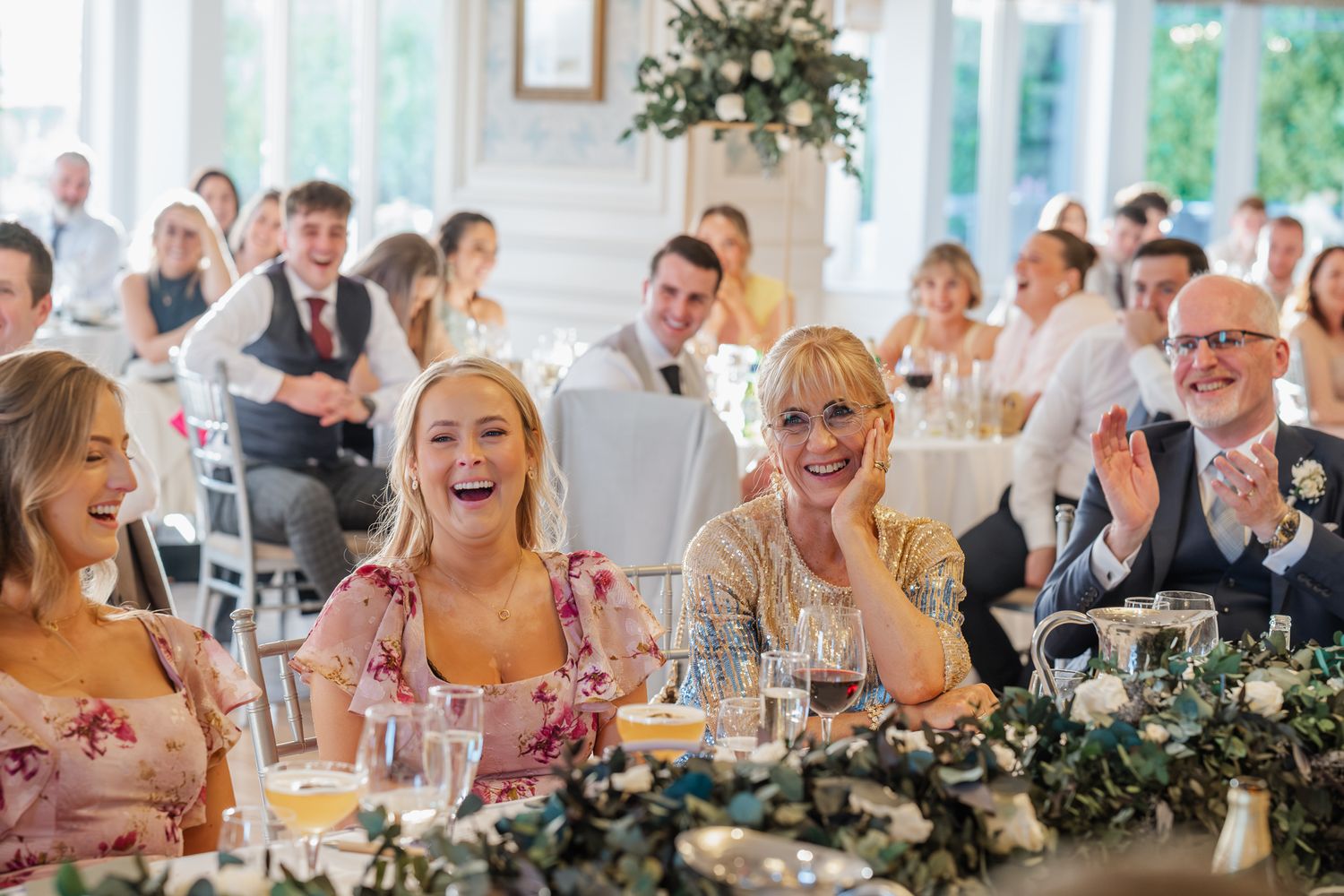 Wedding guests laughing and clapping at a reception, seated at decorated tables in an elegant white venue.