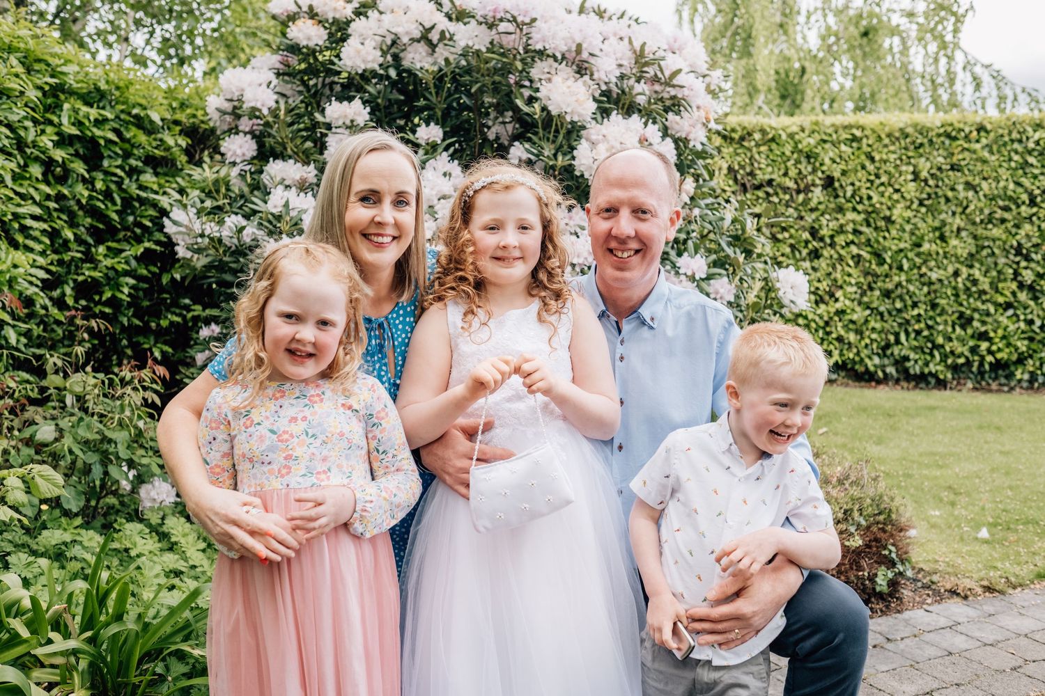Happy family of five posing in a garden with blooming flowers, parents smiling with three young children in formal attire.