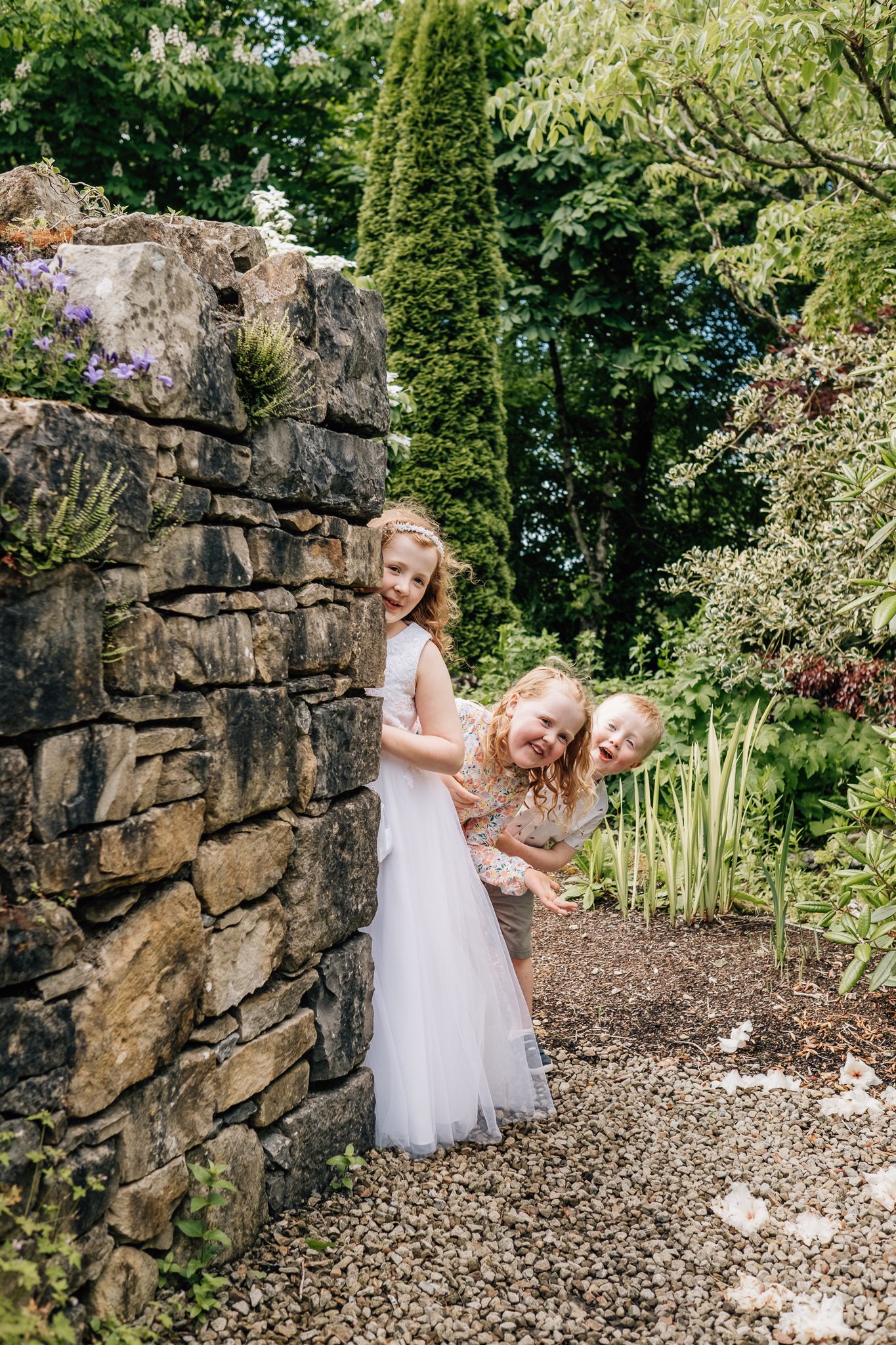 Two young girls in white dresses peeking around a stone wall in a lush garden setting.