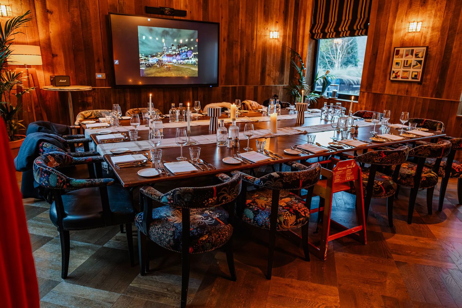 Elegant private dining room with long wooden table, floral chairs, glassware settings, and dual screens in a warm wood-paneled space.