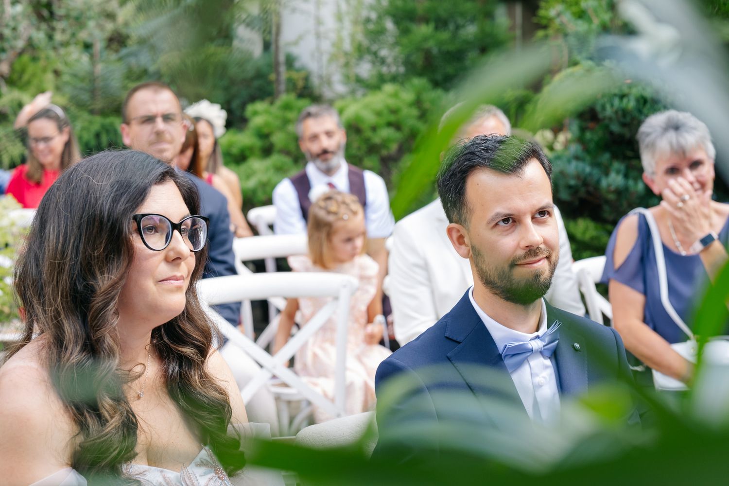 Bride and groom sitting at outdoor wedding ceremony surrounded by guests and lush green foliage.
