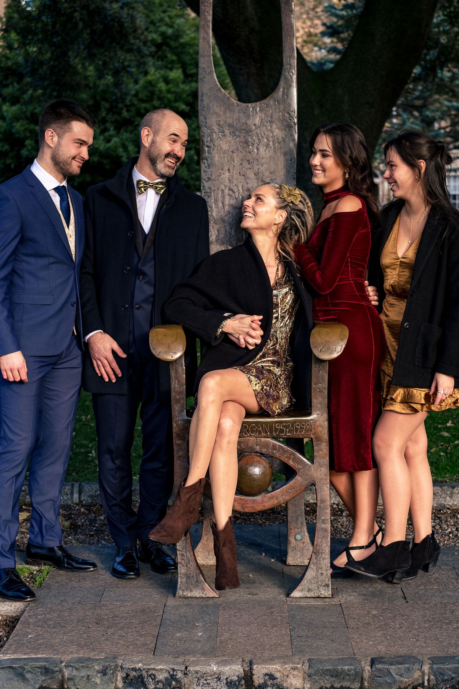 Five elegantly dressed friends laughing and posing around a decorative bench sculpture outdoors.