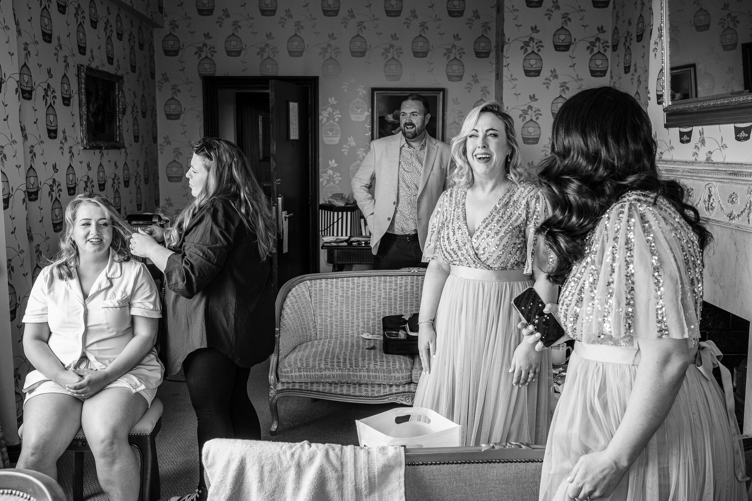 A black and white photograph of wedding preparation moments in a wallpapered room.