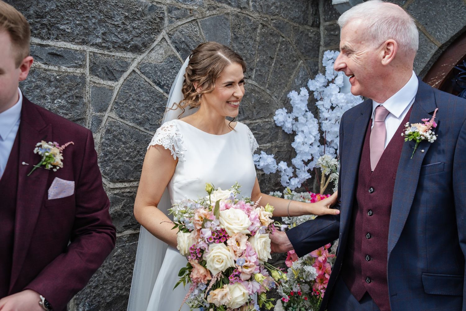 Wedding party in burgundy and navy attire pose together with pink floral bouquets against stone wall.