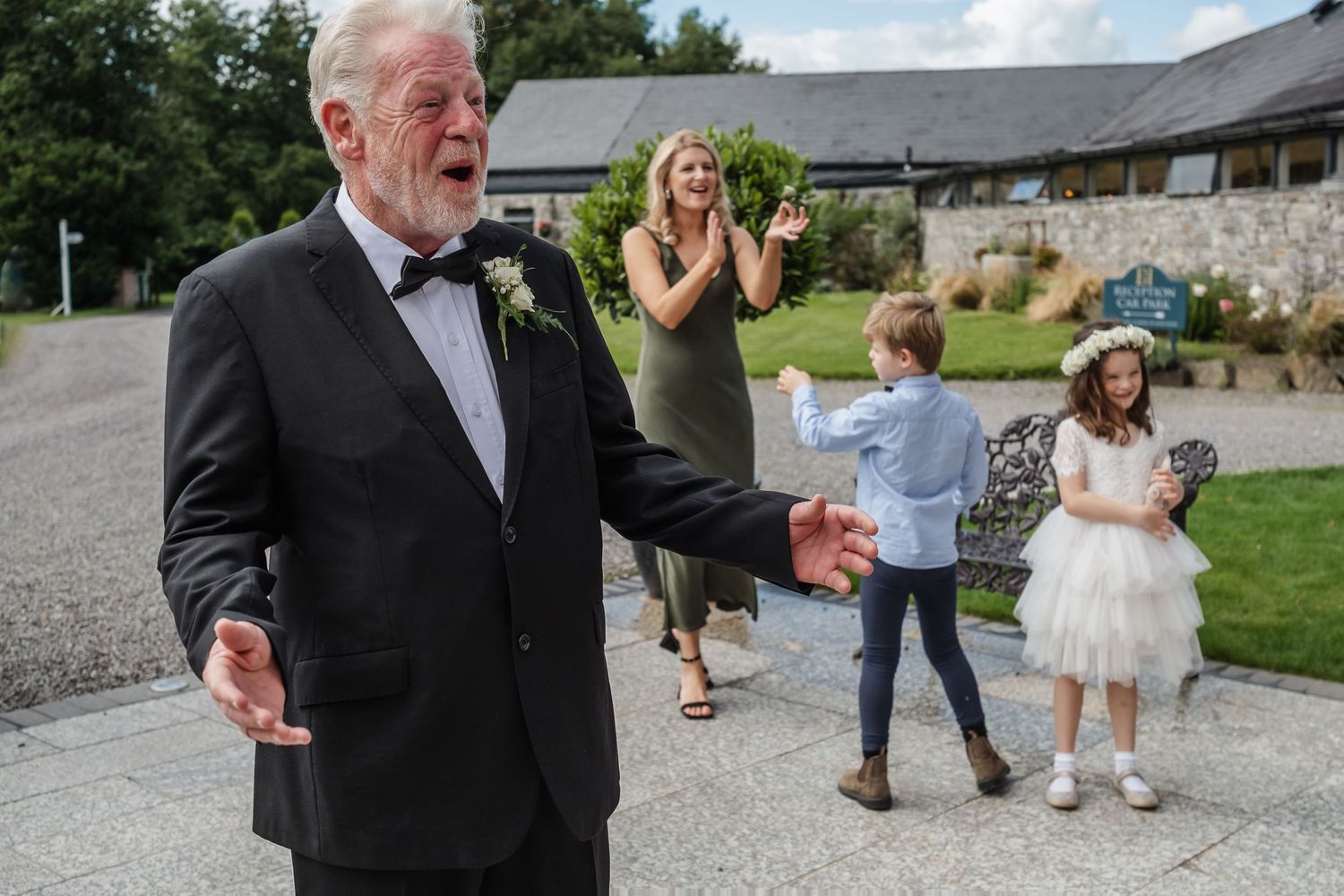 A joyful celebration at a stone country house with people in formal attire dancing on a gravel driveway.