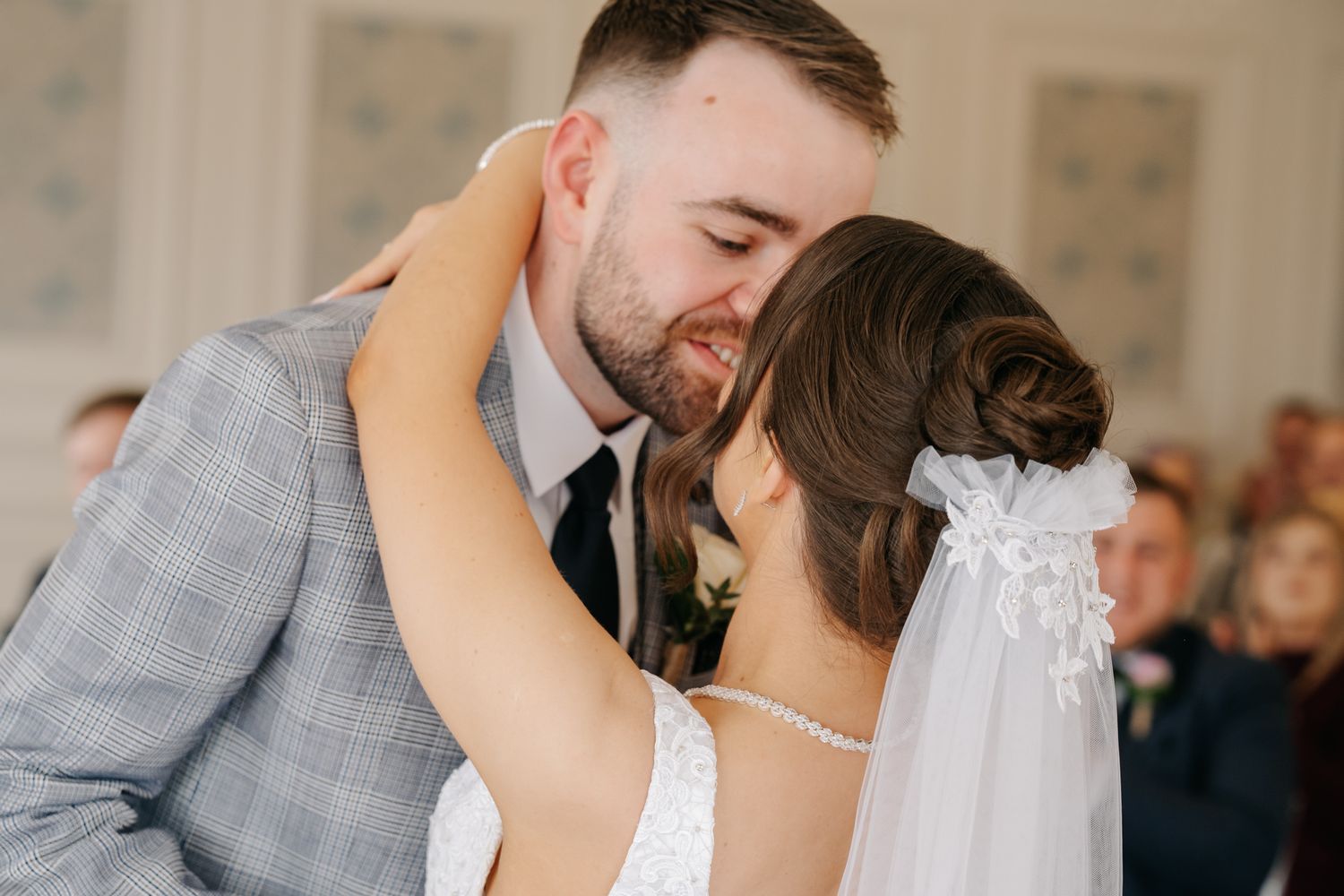 Bride and groom share a tender first dance kiss, bride in white lace gown with veil, groom in grey plaid suit.