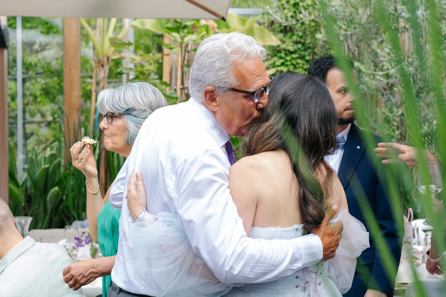Elderly man in white shirt embracing a woman in a strapless dress at an outdoor wedding reception.