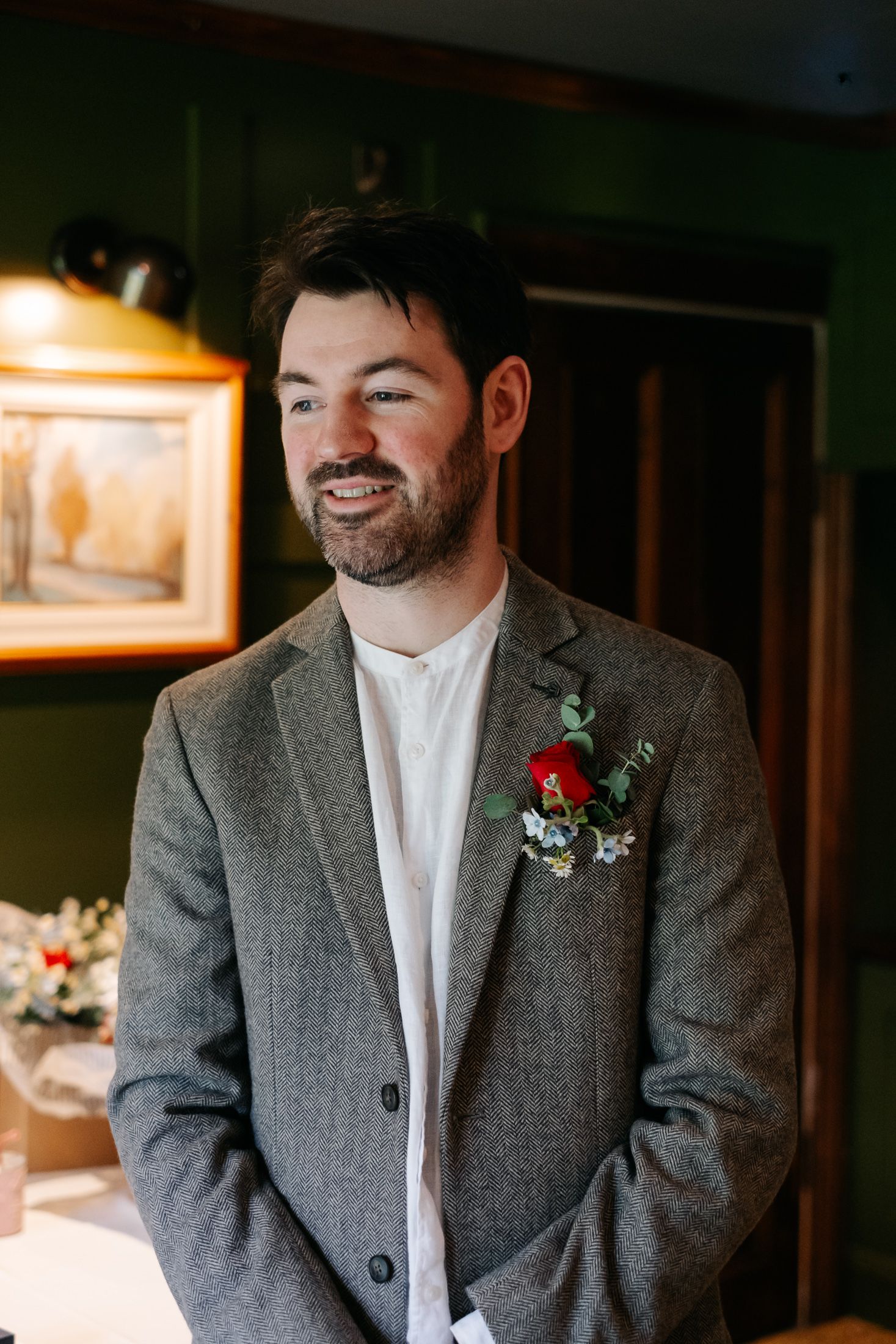 Smiling groom in grey tweed blazer with red rose boutonniere stands in elegant green-walled venue.