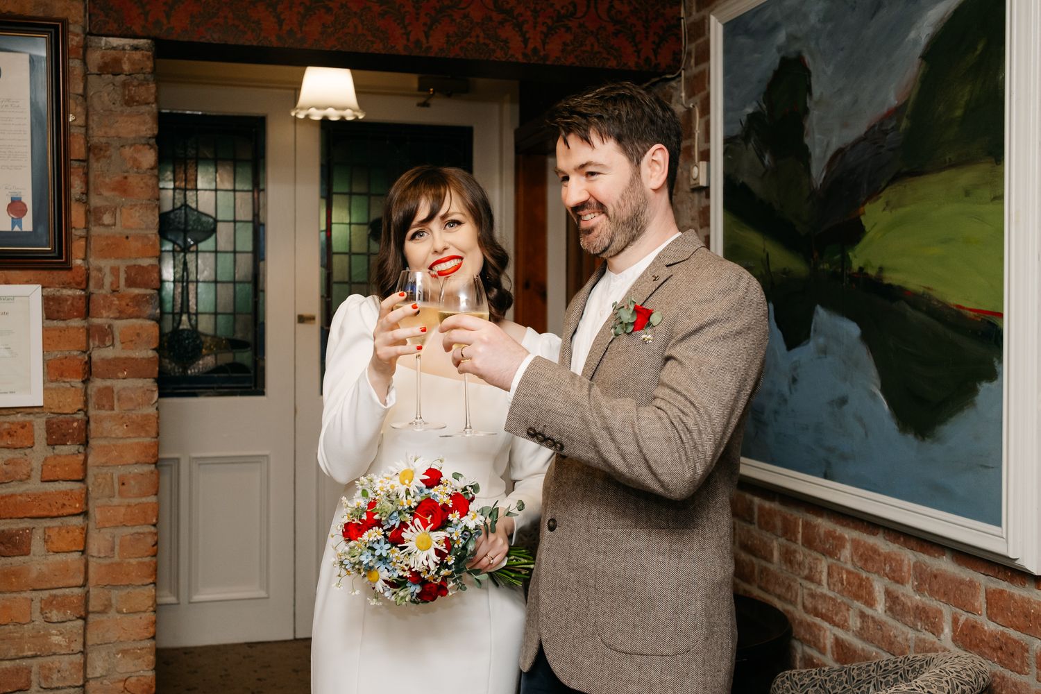 Bride and groom toasting with champagne glasses, bride holding colorful bouquet, standing in rustic brick interior.