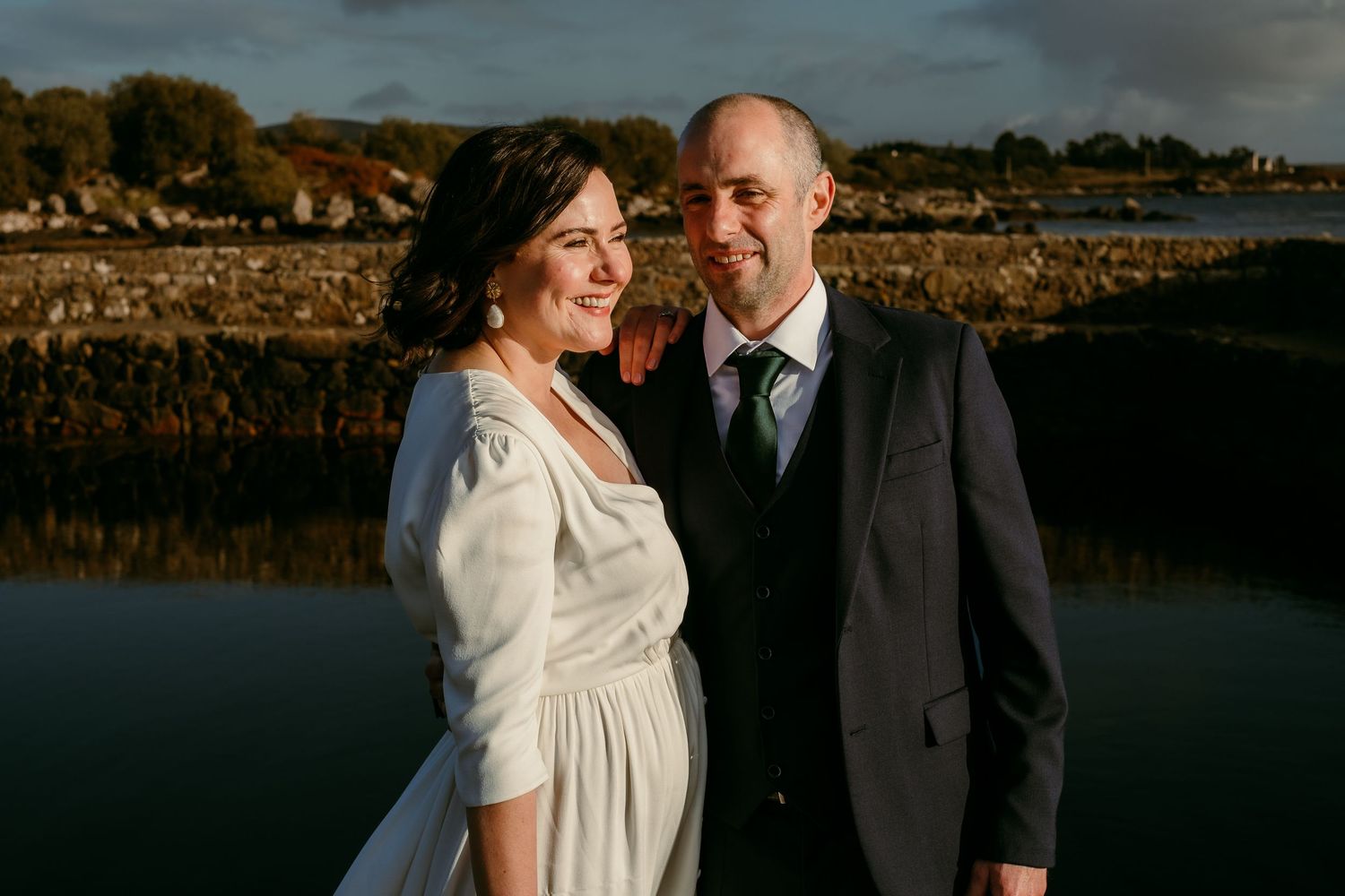Smiling bride and groom standing together by a calm reflective waterway in golden evening light.