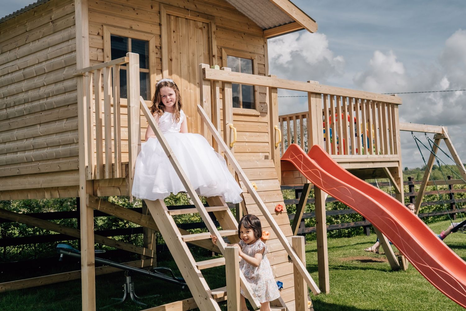 Two girls in white dresses playing on a wooden elevated playhouse with a red slide in a green garden.