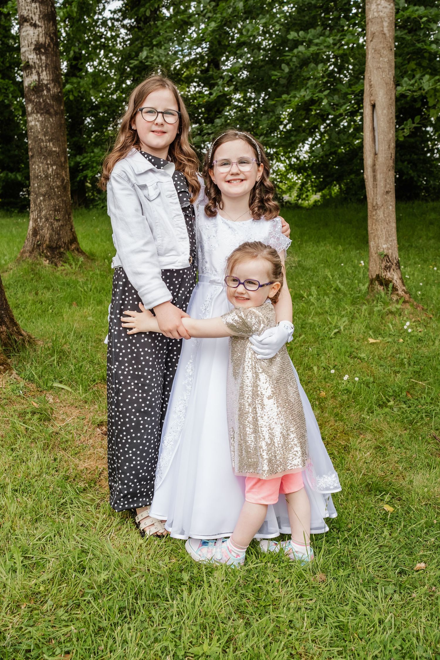 Three young girls in dresses pose together outdoors in a green park setting, with the middle girl wearing a white communion dress.