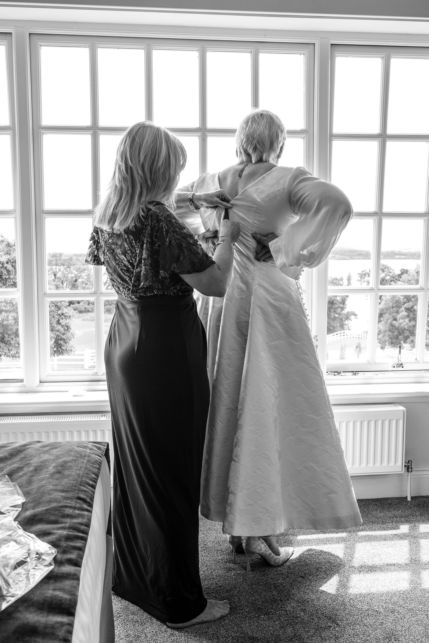 Bride being helped into wedding dress by bridesmaid at sunlit window in black and white photo.