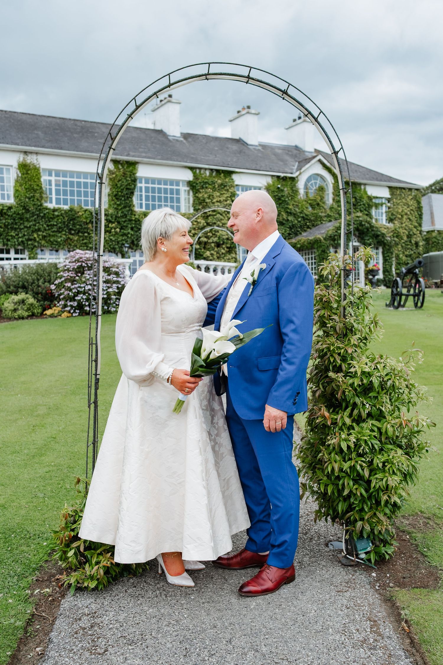 Mature bride and groom in blue suit pose under garden arch at ivy-covered wedding venue.