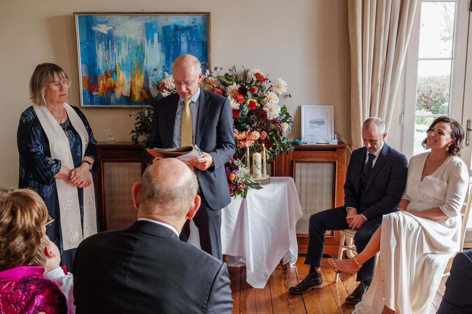 Intimate wedding ceremony with couple seated smiling as officiant reads vows in elegant room with floral arrangement.
